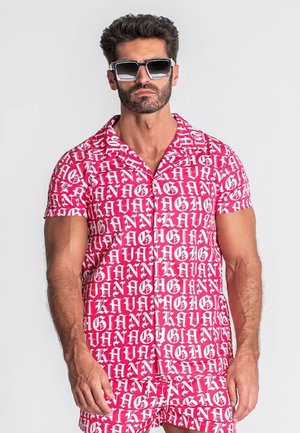 Homme portant une chemise à manches courtes et un short assortis rouges et blancs avec un motif de texte audacieux de style gothique, et de grosses lunettes de soleil carrées noires.