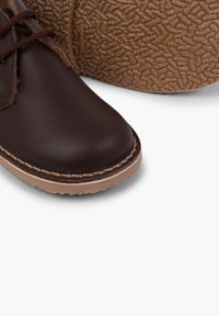 Hnědá kožená bota s kulatou špičkou, hnědými tkaničkami a texturovanou gumovou podrážkou. Viditelné švy podél okraje přidávají na detailech designu.