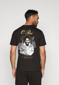 Camiseta negra con un gráfico impreso de un jugador con el texto "O Rei" y "10" en dorado y blanco, con un fondo de diseño texturizado.