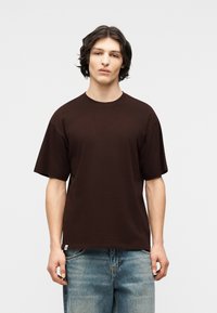 Νέος άνδρας με σκούρα μαλλιά μέχρι τους ώμους, φορώντας ένα απλό υπερμεγέθες σκούρο καφέ μπλουζάκι και ξεθωριασμένο μπλε τζιν, με φόντο ένα γκρι φόντο.