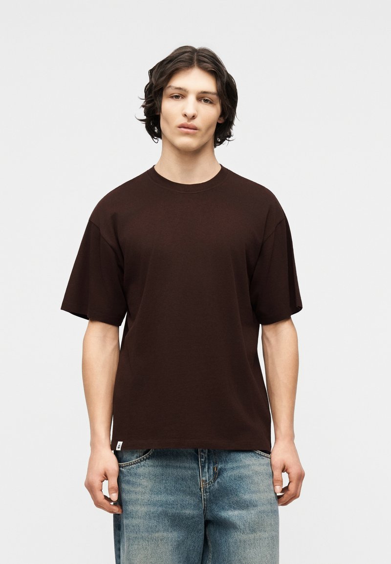 Νέος άνδρας με σκούρα μαλλιά μέχρι τους ώμους, φορώντας ένα απλό υπερμεγέθες σκούρο καφέ μπλουζάκι και ξεθωριασμένο μπλε τζιν, με φόντο ένα γκρι φόντο.