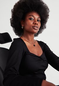 Αυτοπεποίθηση γυναίκα με φυσικό αφρο χτένισμα, που φοράει μαύρο τοπ και χρυσά σκουλαρίκια κρίκους, καθισμένη σε μια μαύρη καρέκλα σε απλό φόντο.