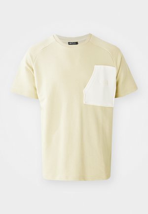 T-shirt beige chiaro con scollo rotondo, maniche raglan corte e una tasca sul petto bianca a contrasto con un logo. Materiale in cotone morbido.