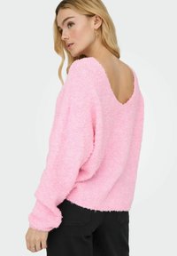 Roza puhasta svetra z odprtim hrbtom, dolgimi rokavi in ohlapnim krojem. Tekstura izgleda mehka in udobna. V kompletu z črnimi hlačami.