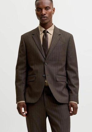 Homme portant un costume marron à fines rayures avec une chemise beige et une cravate marron foncé, debout devant un fond uni.