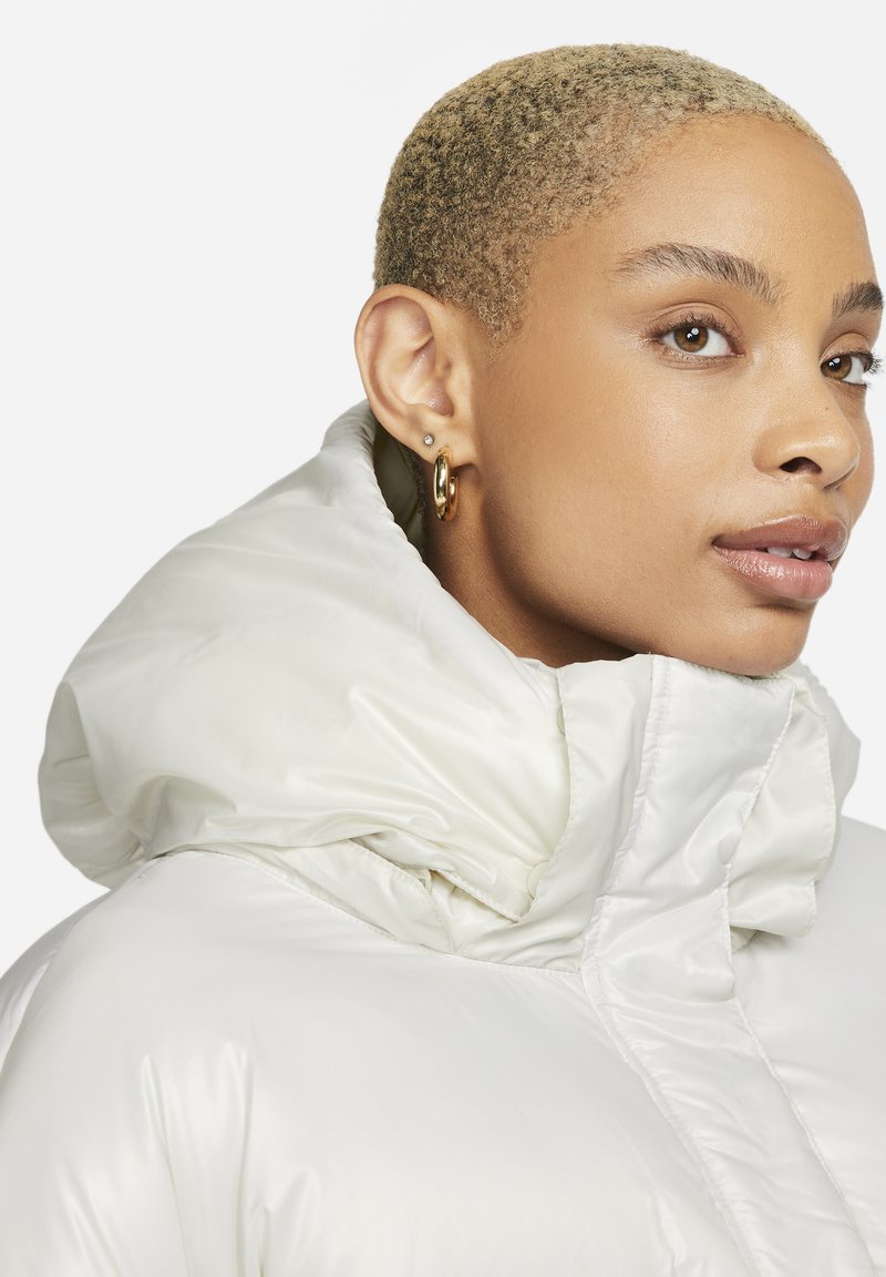 Chaqueta acolchada blanca con una gran capucha de peluche y una textura suave. El diseño está ajustado alrededor de la cara con detalles de pliegue.