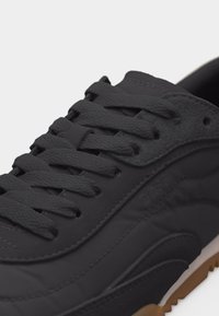 Prim-plan cu un pantof sport negru cu șireturi texturate, panouri din piele întoarsă și material neted, și o margine a talpii din cauciuc gumă.