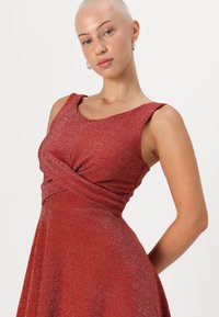 Robe rouge au design sans manches, présentant un détail de taille torsadé. Le tissu a une texture scintillante, créant un effet chatoyant.
