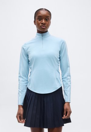 Žena u svijetloplavoj majici s dugim rukavima na patentni zatvarač i crnoj plisiranoj suknji, stoji ispred jednobojne svijetle pozadine.