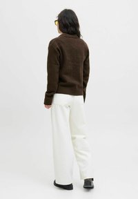 Pull en tricot marron avec un ourlet côtelé, associé à un pantalon large blanc et des chaussures noires. Le modèle est vu de dos.