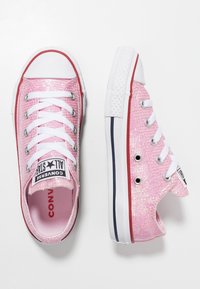 Różowe lśniące trampki na płóciennym materiale z białymi sznurówkami i gumowymi podeszwami. Posiadają białą osłonę palców oraz czerwone akcenty wzdłuż podeszwy i oczek na sznurówki.