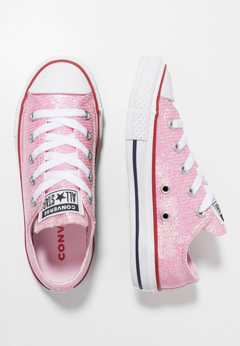 Różowe lśniące trampki na płóciennym materiale z białymi sznurówkami i gumowymi podeszwami. Posiadają białą osłonę palców oraz czerwone akcenty wzdłuż podeszwy i oczek na sznurówki.