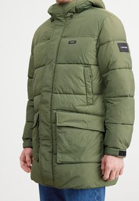 Mand iført en olivengrøn, polstret Calvin Klein vinterjakke med flere lommer og hætte, parret med blå jeans.