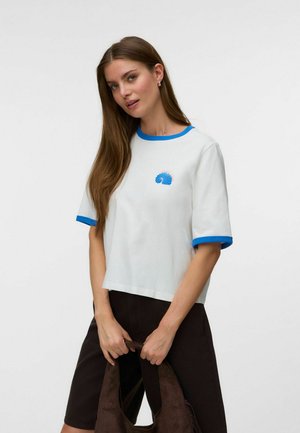 Kvinde med langt brunt hår har hvid t-shirt med blå kant og skaldyrsdesign, sorte shorts, holder en mørkebrun taske mod en ensfarvet baggrund.