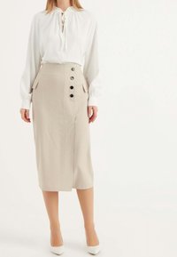 Jupe midi beige à rayures avec fente à l'avant, poches latérales et boutons noirs. Associée à une blouse blanche avec un col à nouer.