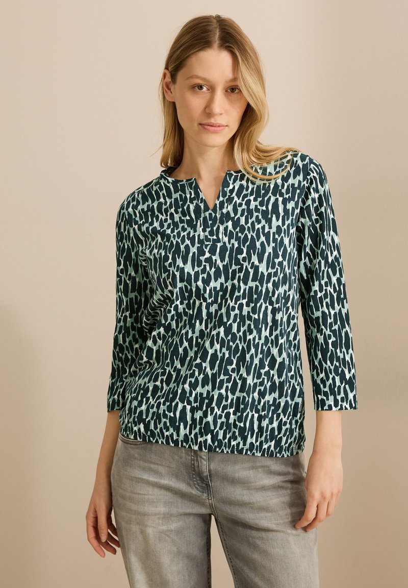 Turkos och svart mönstrad blus med V-hals, trekvartslång ärm och en lös passform, kombinerad med ljusgrå denim.