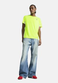 Fluorescencyjna żółta koszulka z krótkimi rękawami, zestawiona z jasnoniebieskimi spodniami typu wide-leg i czerwonymi butami wsuwanymi. Prosty krój, bez wzorów.