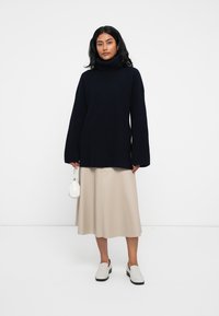 Navy rebrast pulover s turtleneck ovratnikom in dolgimi rokavi, v kombinaciji s kremno A-linijo krila in belimi natikači, drži majhno belo torbico.