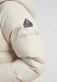 Beige pufjakke sektion med quiltet design, prydet med et sort trekantet logo-patch med teksten "JACKIT" på ærmet.