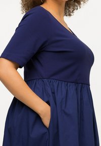 Marinblå klänning med korta ärmar, V-ringning och figurnära överdel. Utrullad kjol med rynkad textur, har sidofickor.