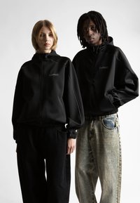 Két ember áll egymás mellett, fekete zipzáras kapucnis pulóverben, amelyeken a "wasted" felirat látható, az egyik fekete nadrágot, a másik pedig kifakult kék farmert visel.