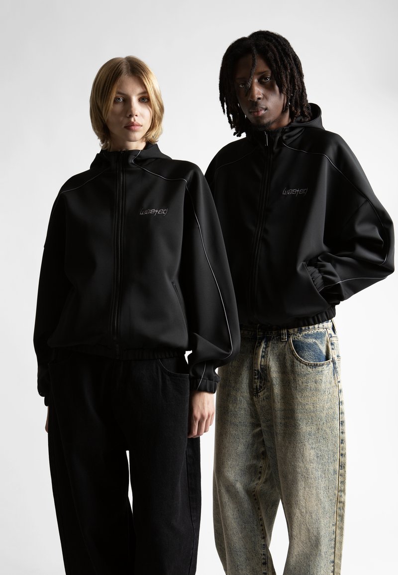 Zwei Personen stehen nebeneinander und tragen schwarze Reißverschluss-Hoodies mit dem Text "wasted". Eine trägt schwarze Hosen und die andere ausgebleichte blaue Jeans.