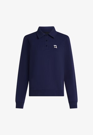 Tumši zilas krāsas garām piedurknēm polo krekls, izgatavots no mīksta auduma, ar trīs pogām un mazu zīmola logo kreisajā krūšu daļā.