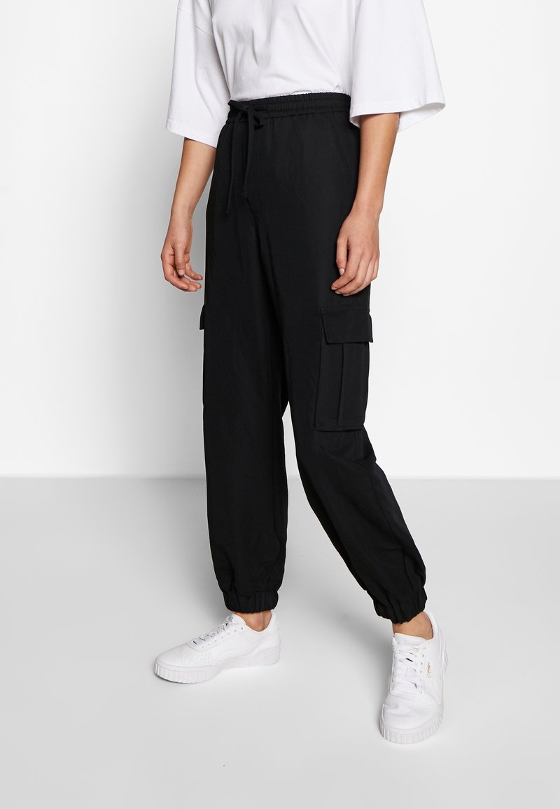 Personne portant un pantalon cargo jogger noir avec des poignets élastiques, des baskets blanches et une chemise blanche ample, debout devant un fond uni.
