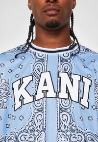 Lyseblå trøye med paisley-mønster og svart-hvite detaljer. Fet "KANI"-tekst fremhevet i hvitt.