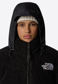 Czarna polarowa kurtka z zamkiem błyskawicznym, o fakturowej powierzchni i regulowanym kapturze. Wyraźne szare logo na klatce piersiowej.