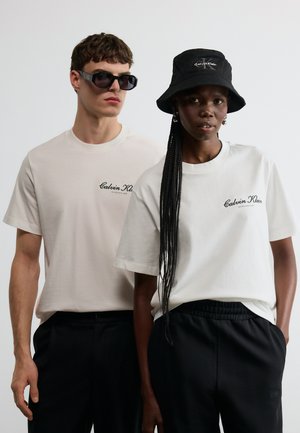 Zwei Models tragen weiße Calvin Klein T-Shirts und schwarze Hosen, eines mit Sonnenbrille, das andere mit einem schwarzen Calvin Klein Bucket Hat.