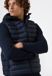 Młody mężczyzna z kręconymi włosami, ubrany w granatowy puchowy kamizelkę z kapturem oraz koszulę z długim rękawem, z założonymi rękami, patrzy w lewo.