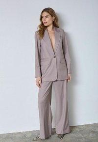 Svetlosivé oblečenie na mieru so sakom s hlbokým výstrihom do V a širokými nohavicami. Jemná látka s elegantnými líniami a zapínaním na jeden gombík.