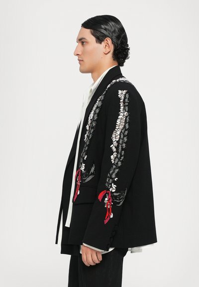 Schwarzer Blazer mit aufwendiger silberner und roter Stickerei, weiten Ärmeln und dekorativen Knöpfen. Über einem Hemd mit Kragen getragen.