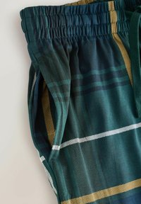 Tummanvihreät ja laivastonsiniset ruudulliset kangasshortsit, joissa on joustava vyötärönauha ja sivutasku, päällä vaalealla taustalla keltaisia ja valkoisia raitoja.