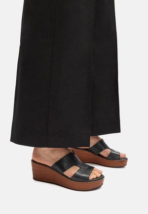 Noge v črnih usnjenih platformah s smešnimi rjavimi teksturiranimi podplati, delno pokrite z dolgimi črnimi širokimi hlačami.