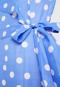 Un tissu à pois bleus présente de grands cercles blancs. Un nœud noué à la taille accentue le design. Le matériau semble léger et fluide.