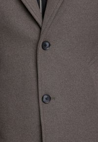 Cappotto in lana marrone con una consistenza morbida, due bottoni neri e un design sartoriale con una leggera lucentezza. Dettagli della chiusura con cerniera e bottone visibili.