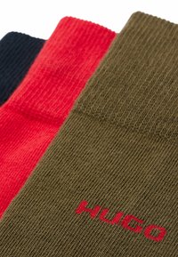 Trois paires de chaussettes : une marine, une rouge et une vert olive avec une texture côtelée et le logo "HUGO" en rouge sur la paire olive.
