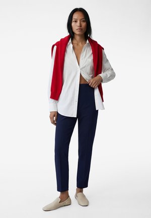 Kvinde stående iført en hvid skjorte med knapper, delvist åben, marineblå bukser, beige slip-on sko og en rød sweater hængende over skuldrene.