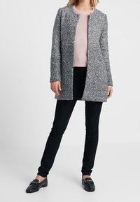 Cappotto oversize grigio in tessuto texturizzato, con apertura frontale e scollo rotondo, indossato sopra un maglione rosa e pantaloni skinny neri. Mocassini neri completano il look.
