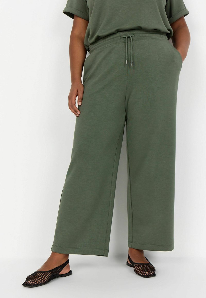 Weite Sweatpants in Olivgrün, aus weichem Material. Mit elastischem Bund und Kordelzug sowie seitlichen Taschen. Kombiniert mit schwarzen Mesh-Ballerinas.