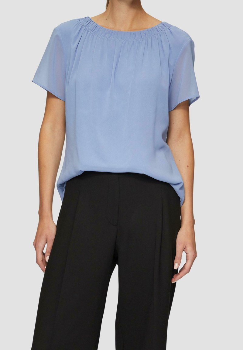 Femme portant un chemisier bleu clair à manches courtes avec un col froncé, rentrée dans un pantalon noir taille haute à jambes larges sur fond gris.
