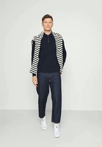 Camisa polo azul-marinho combinada com calças de ganga escuras e sapatilhas brancas. Suéter listrado pousado sobre os ombros. Tecido suave, design casual.