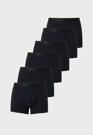 Homme athlétique à la peau foncée, debout pieds nus, bras croisés, portant un boxer noir ajusté sur un fond clair uni.