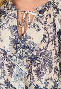 Blumenbluse aus leichtem Stoff mit blauen Blumenmustern, einem Rüschensaum am Ausschnitt und einem gebundenen Detail an der Vorderseite mit einer offenen V-Form.