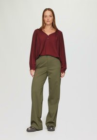 Blouse bordeaux à manches longues avec un décolleté en V et des boutons, associée à un pantalon taille haute vert olive et des chaussures à plateforme sombres, modèle debout.
