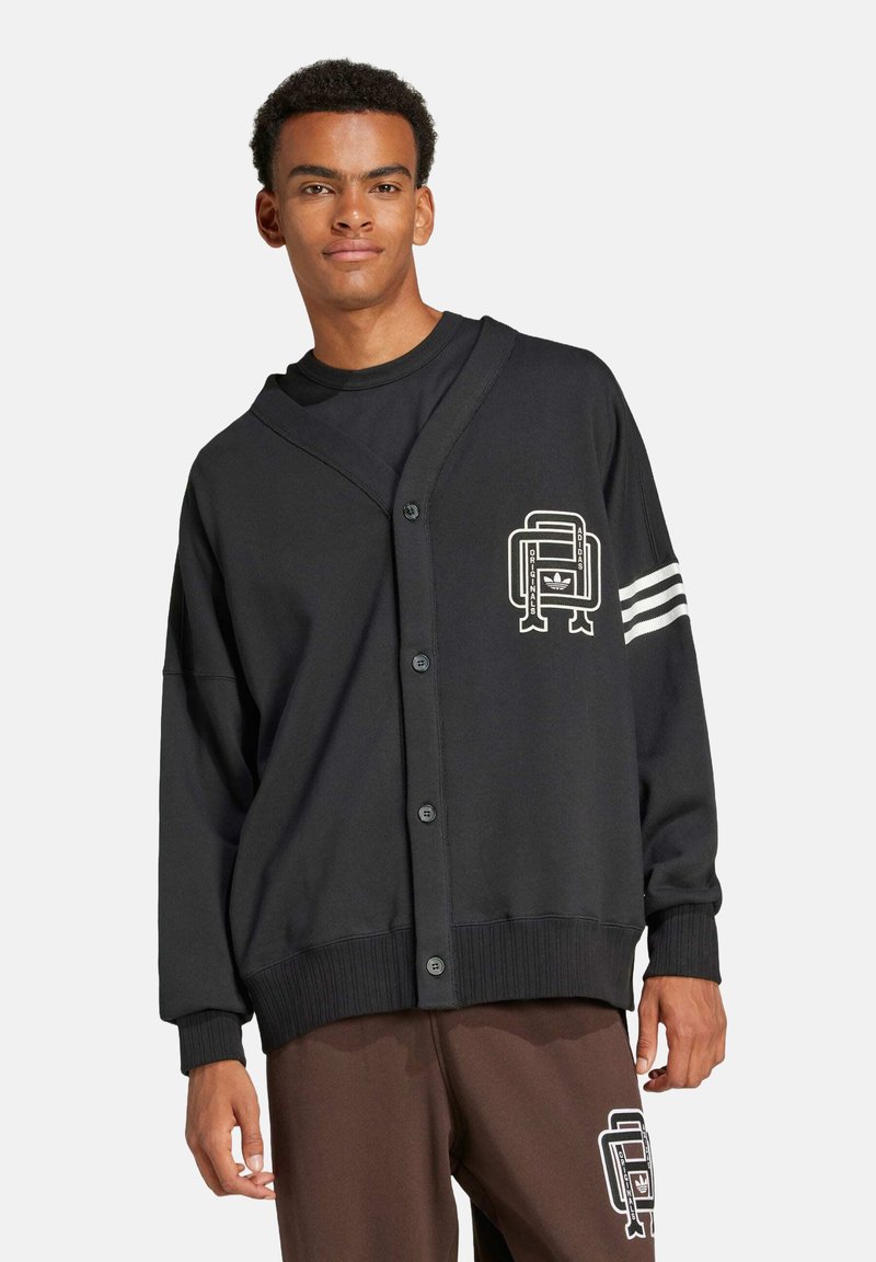 Sort cardigan med knaplukning, ribbede manchetter og oversize pasform. Har et grafisk motiv på venstre bryst og hvide striber på ærmet.