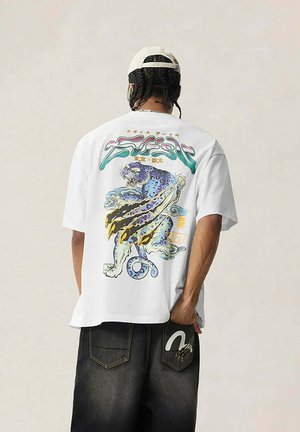 Persona con capelli intrecciati che indossa un berretto bianco, una T-shirt bianca con una grafica colorata di una pantera ruggente e jeans neri con un disegno sulla tasca posteriore.