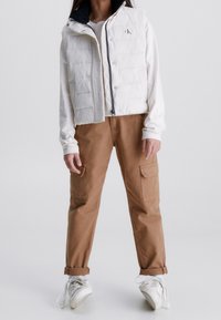 Weißer Puffer-Weste mit durchgehendem Reissverschluss, über einem weißen Langarmshirt getragen. Braune Cargo-Hosen mit aufgesetzten Taschen und weißen Turnschuhen.